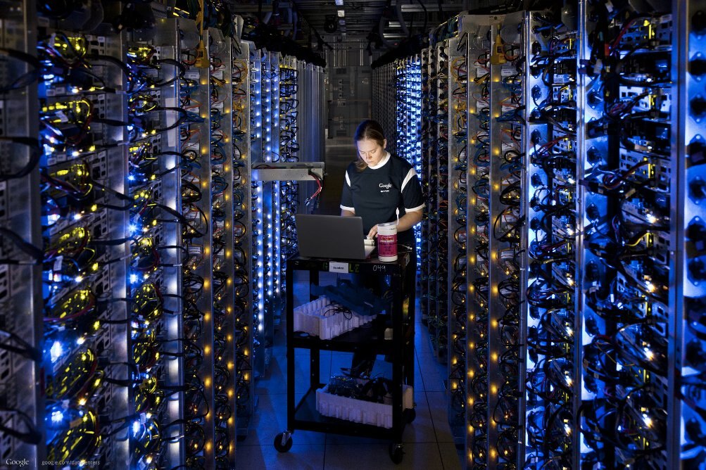 A worker in a data center operating a laptop surrounded by server racks illuminated with blue lights.