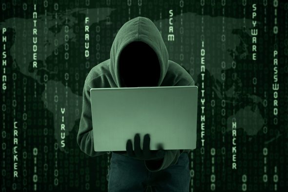 A figure in a hoodie using a laptop,set against a digital background with words related to cybercrime and security.