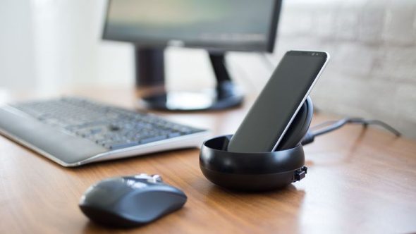 A modern workspace featuring a smartphone in a black docking station,next to a computer monitor,keyboard,and a mouse.