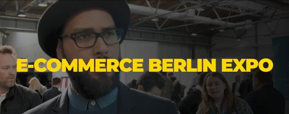 A man in a black hat and glasses at the E-Commerce Berlin Expo with a crowd in the background.