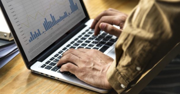 A person typing on a laptop with financial graphs displayed on the screen.