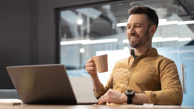 A man sitting at a desk,smiling and holding a cup of coffee while looking at his laptop.