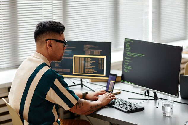 A man seated at a desk working on a computer surrounded by multiple screens displaying code and programming interfaces.