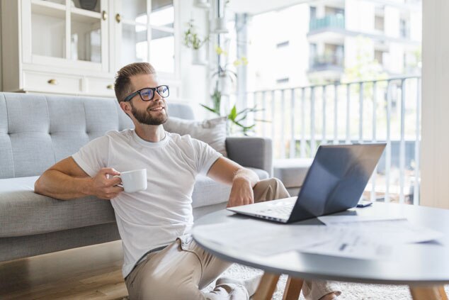 A man sitting on the floor next to a sofa,enjoying a cup of coffee while working on a laptop.