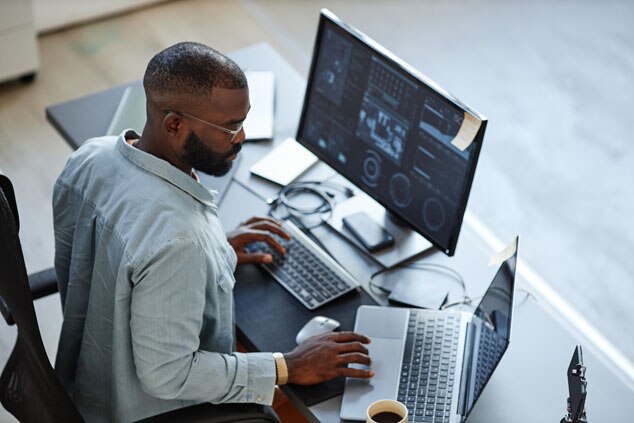 A man working at a desk with multiple computer screens,focused on his tasks,with a cup of coffee nearby.