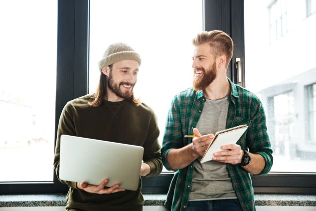 Two young men engaging in a discussion while one holds a laptop and the other takes notes.